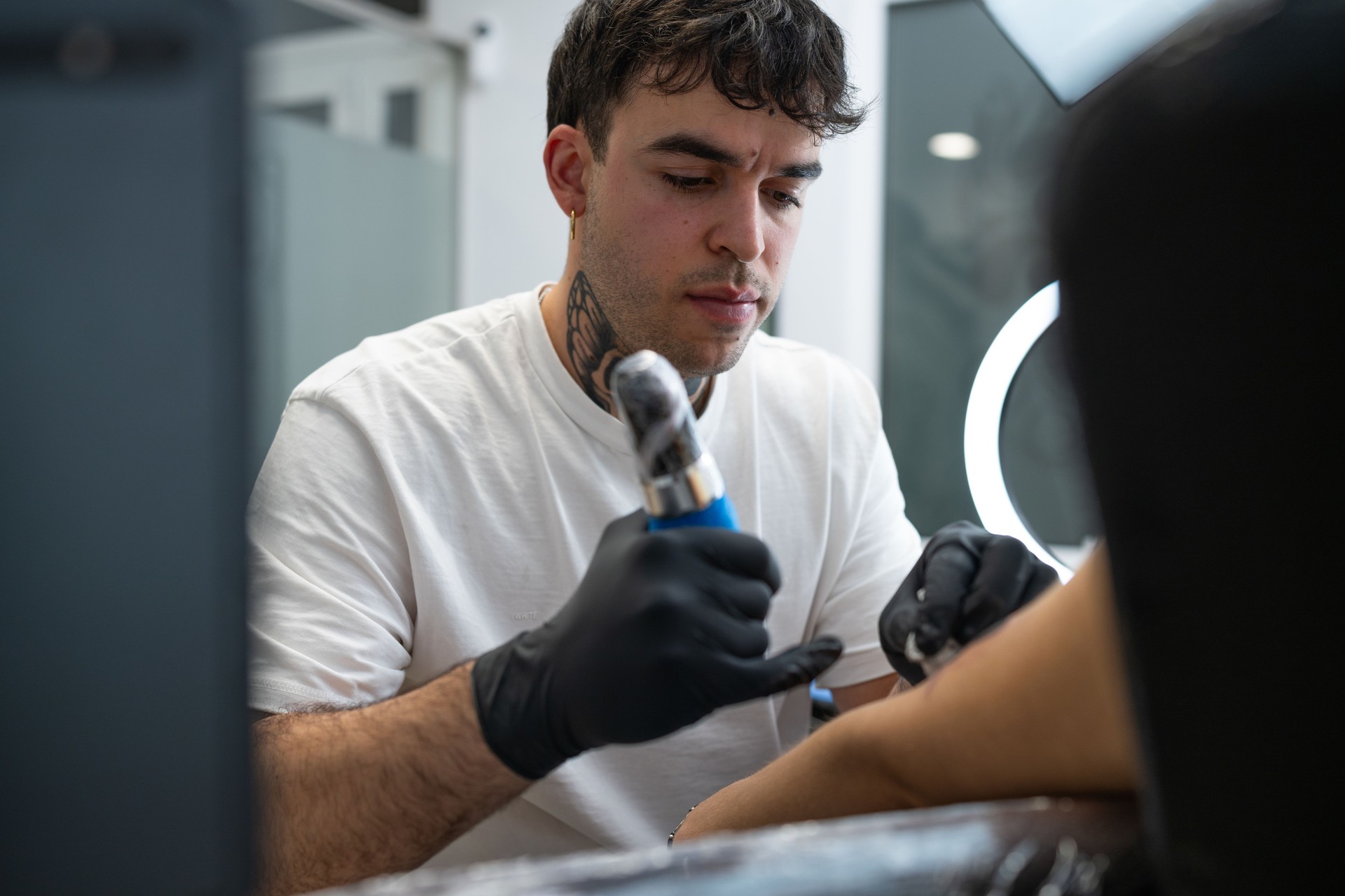 Tattoo professional wearing black gloves carefully inking detailed design onto client's arm, working with precision inside modern studio environment
